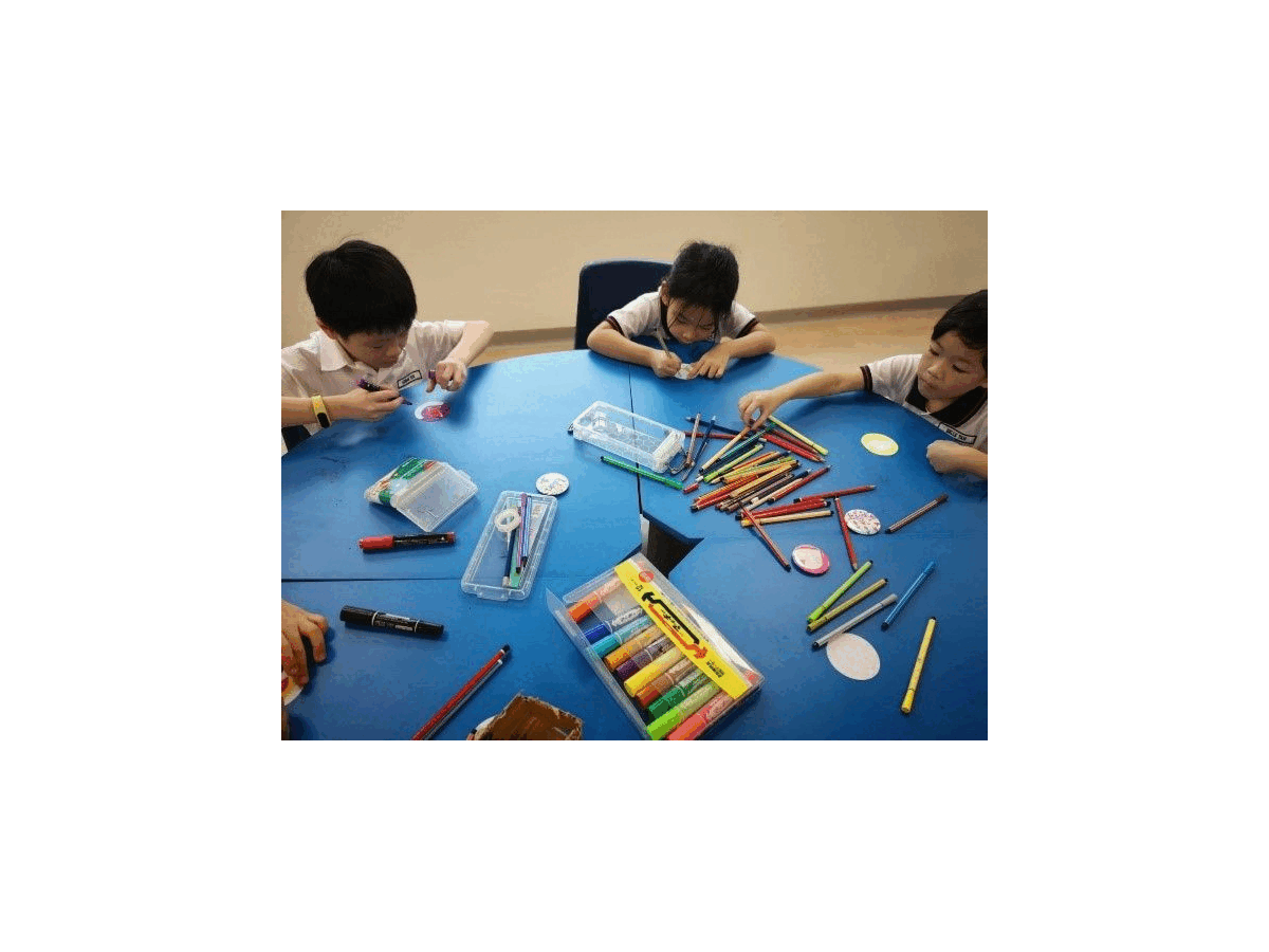 Three children sit around a blue round table covered with colored pencils, markers, and crayons doing a craft activity.